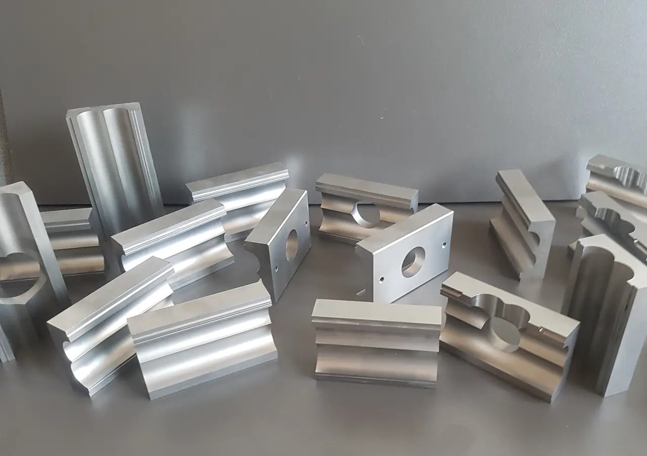 Surowe, aluminiowe elementy obrobione w Brzóze, z widocznymi śladami frezowania i otworami, ułożone na szarym tle.