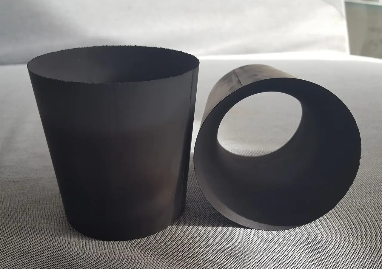 Dwa ciemne, metalowe elementy: stożkowy cylinder z nierównym brzegiem i krótki odcinek rury, oba na szarym tle.