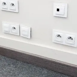 Białe gniazdka elektryczne i włączniki światła zamontowane na kremowej ścianie nad szarą listwą przypodłogową i szarym dywanem, widoczny delikatny pęknięcie na ścianie.