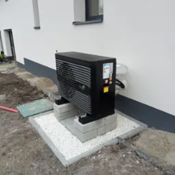 Czarna jednostka zewnętrzna pompy ciepła marki FOX Air, zamontowana na betonowych bloczkach i otoczona białym żwirem, przy białej ścianie budynku z szarym cokołem i oknem. Widoczne rury w izolacji.