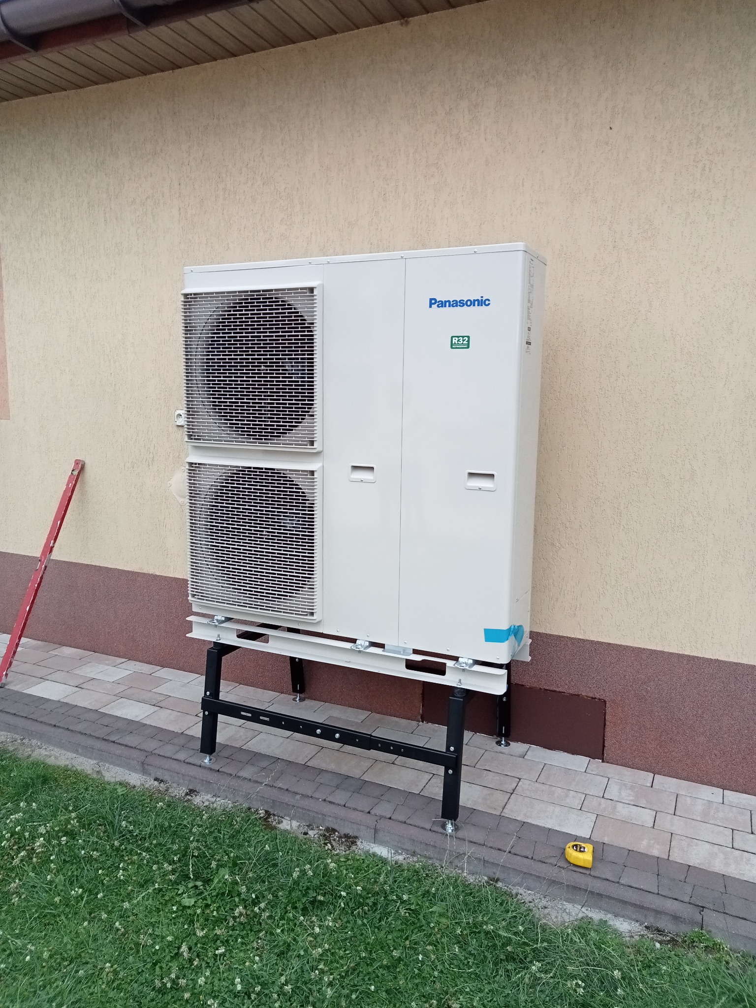 Zewnętrzna jednostka klimatyzacji Panasonic R32 zamontowana na metalowym stelażu przy elewacji budynku, widoczna poziomica i miarka zwijana.