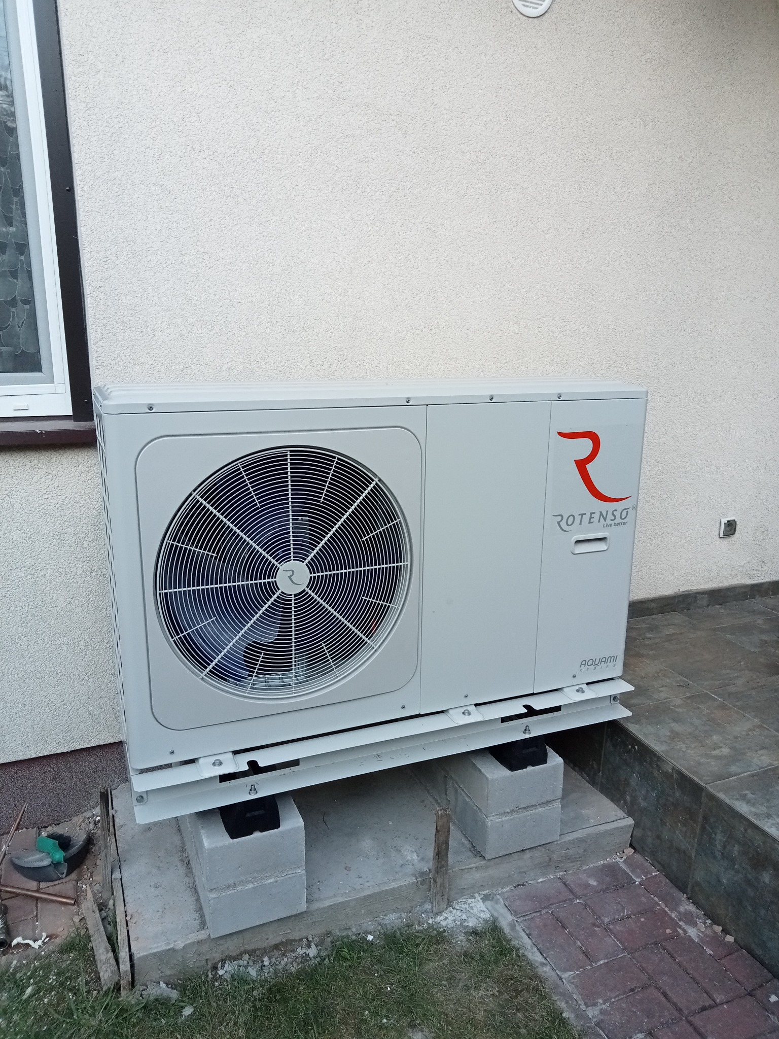 Zewnętrzna jednostka grzewcza Rotenso Aquami Series, zamontowana na betonowych bloczkach przy ścianie budynku z widocznym oknem i fragmentem chodnika z kostki.