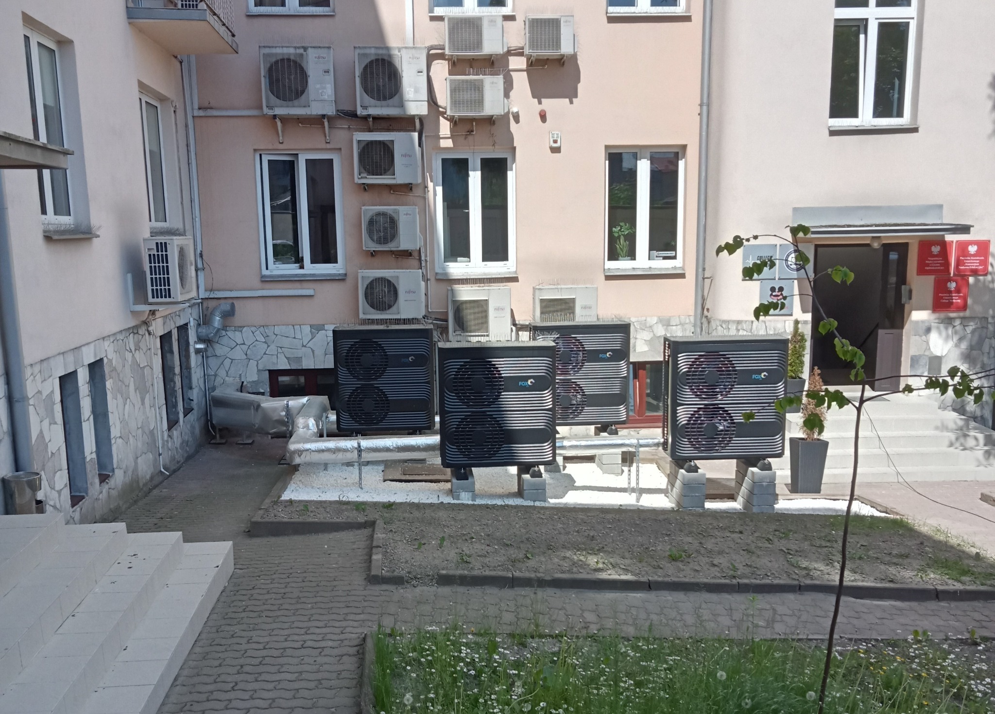 Zewnętrzne jednostki grzewcze marki FOX zamontowane przy budynku z licznymi klimatyzatorami na ścianie, widok z poziomu trawnika.