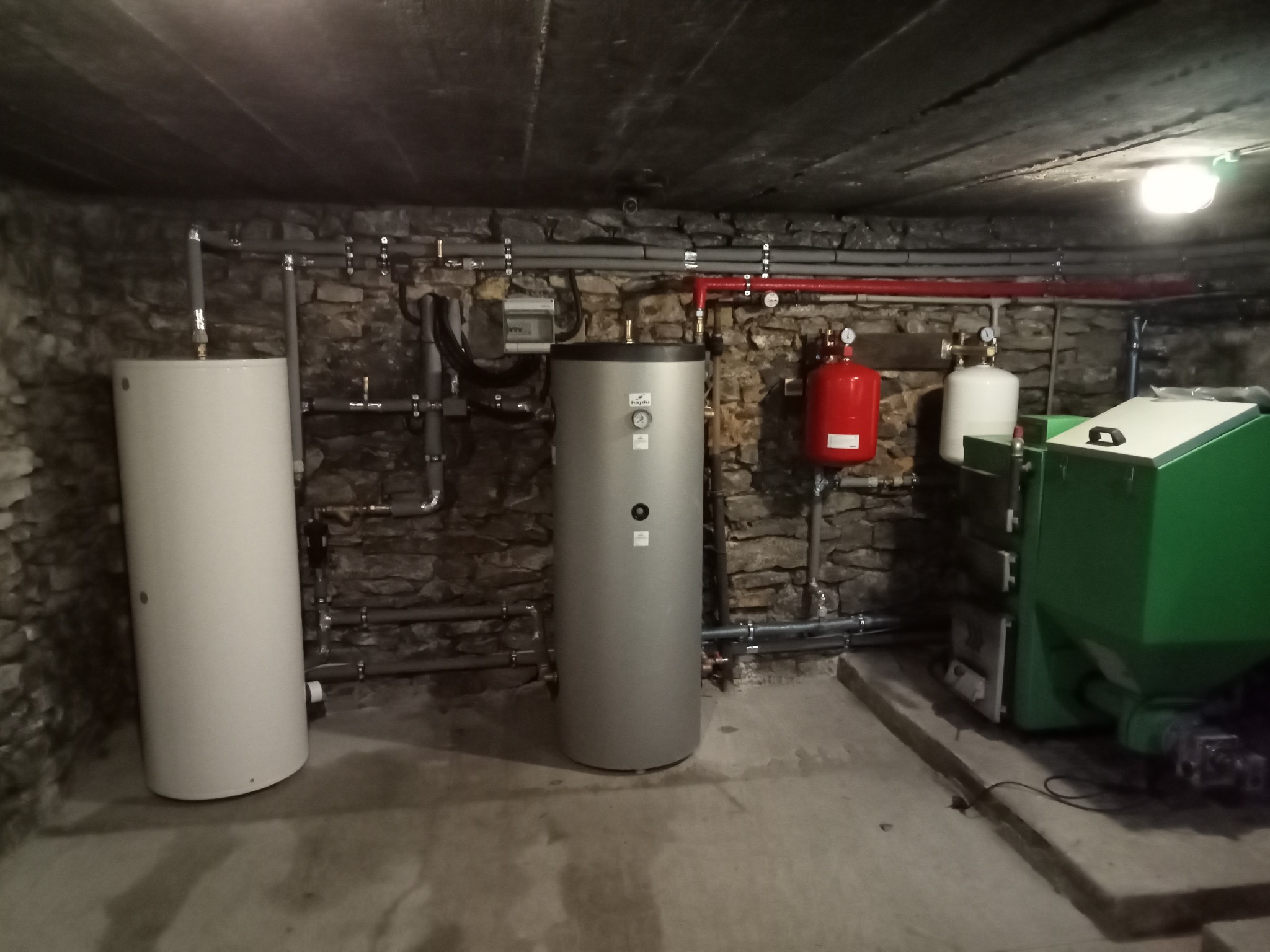 Instalacja grzewcza w piwnicy z kamiennymi ścianami, widoczne dwa zbiorniki akumulacyjne, zasobnik ciepłej wody użytkowej marki Hajdu oraz piec na paliwo stałe z podajnikiem.