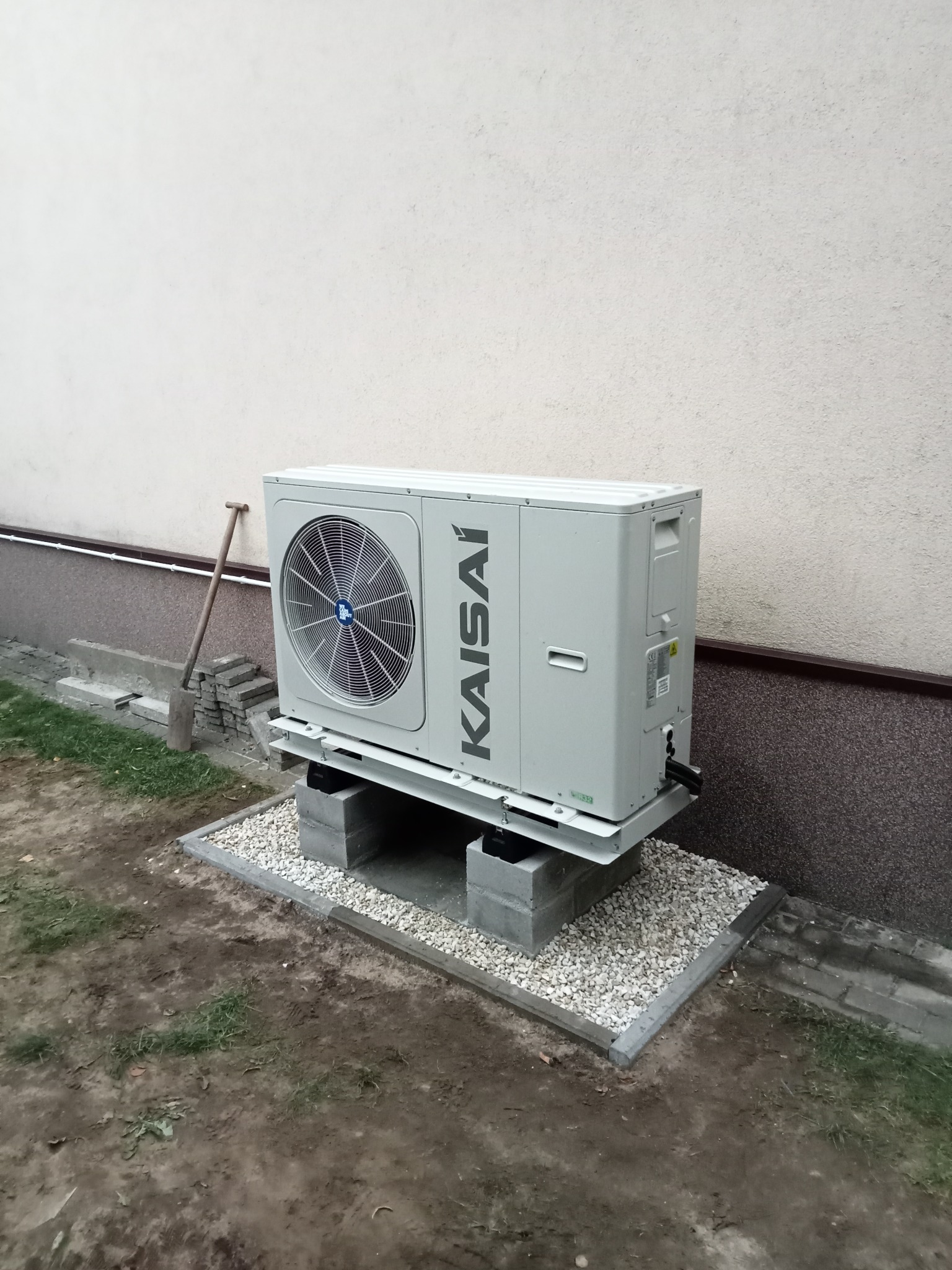 Zewnętrzna jednostka klimatyzacyjna marki Kaisai, zamontowana na betonowych bloczkach i platformie, z widocznymi instalacjami, ustawiona na żwirowym podłożu przy ścianie budynku. Obok leży szpadel.