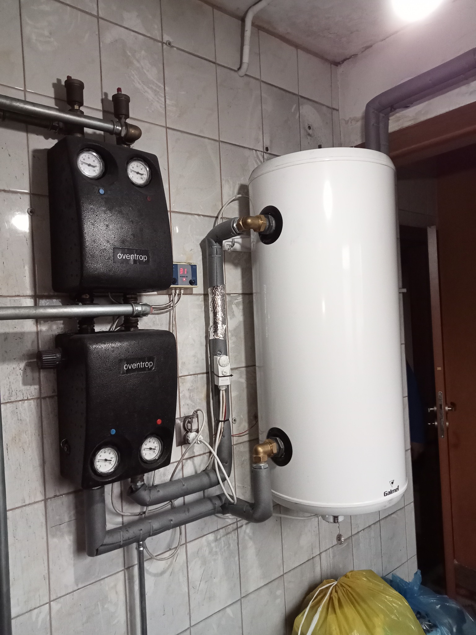 Bojler Galmet podłączony do dwóch regulatorów temperatury Oventrop na ścianie z płytek, widoczne rury i instalacja.