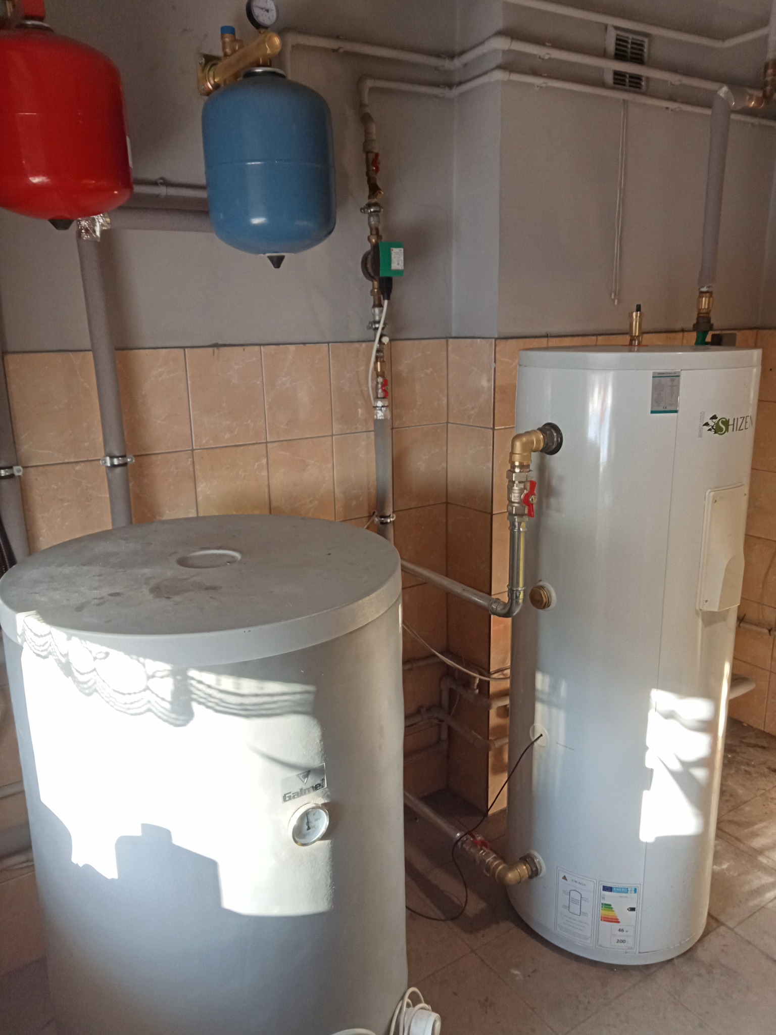 Dwa białe zbiorniki na wodę (Galmet i SHIZEN) z rurami i zaworami, czerwony i niebieski zbiornik wyrównawczy na ścianie z brązowymi płytkami, widoczne manometry i etykiety energetyczne.