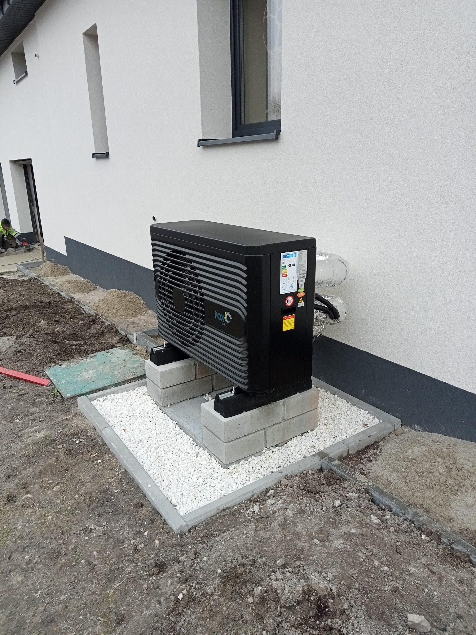 Czarna jednostka zewnętrzna pompy ciepła marki FOX Air, zamontowana na betonowych bloczkach i otoczona białym żwirem, przy białej ścianie budynku z szarym cokołem i oknem. Widoczne rury w izolacji.