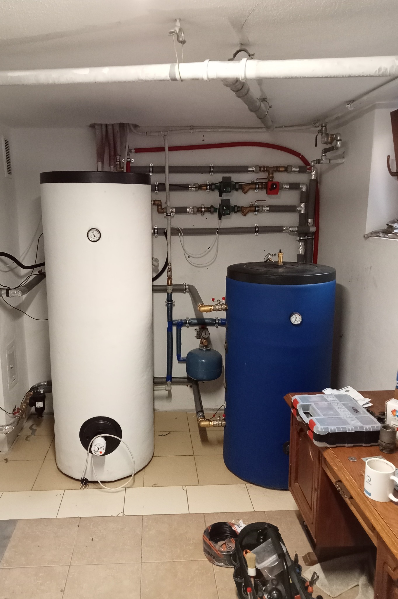 Biały i niebieski zasobnik ciepłej wody w piwnicy, widoczne rury i zawory, skrzynka z narzędziami na drewnianej szafce obok.