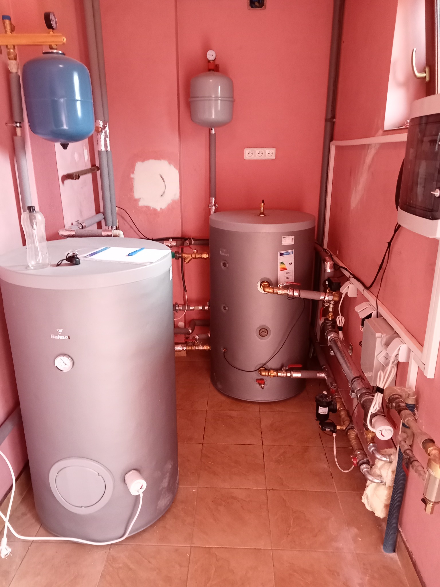 Dwa szare zbiorniki akumulacyjne w kotłowni z różowymi ścianami, widoczne liczne rury i zawory, na jednym ze zbiorników leży notatnik i długopis.