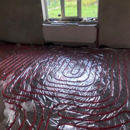 Berben - Czerwone rurki ogrzewania podłogowego ułożone na srebrnej folii izolacyjnej w surowym pomieszczeniu z grzejnikiem pod oknem.