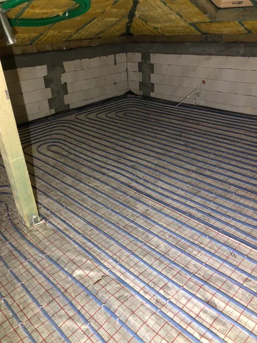 Niebieskie rurki ogrzewania podłogowego ułożone na siatce w surowym wnętrzu poddasza z ociepleniem stropu i ścianami z betonu komórkowego.