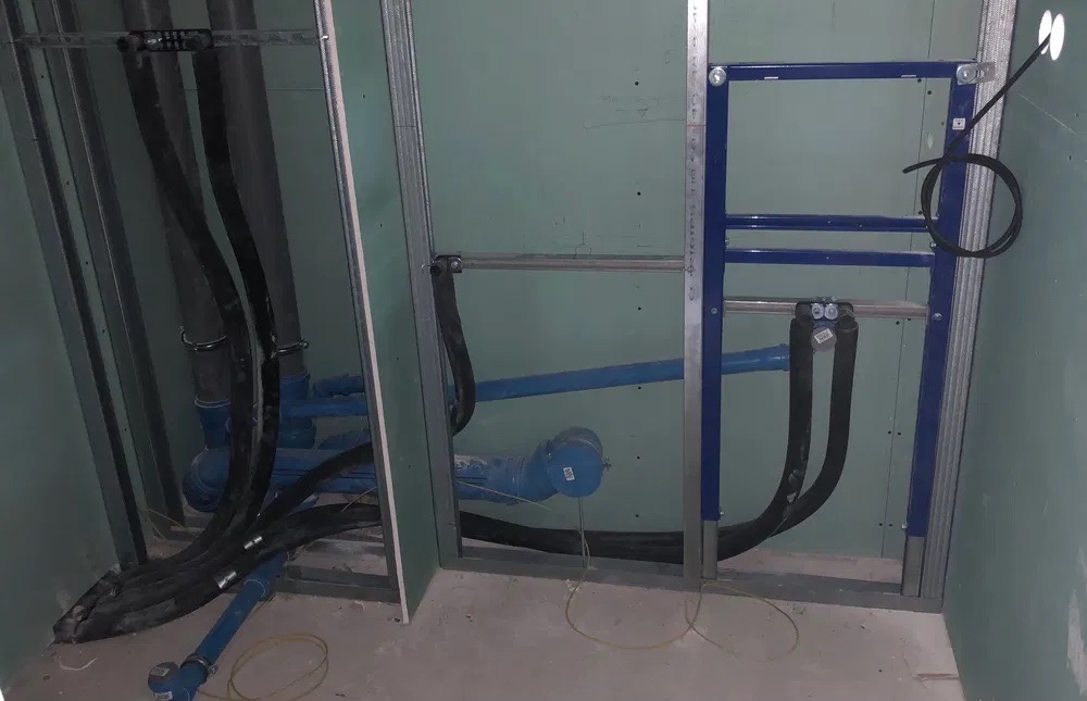 Surowe wnętrze łazienki w trakcie instalacji: widoczne niebieskie rury wodne, czarne rury kanalizacyjne, metalowa rama stelaża podtynkowego WC, płyty gipsowo-kartonowe na ścianach.