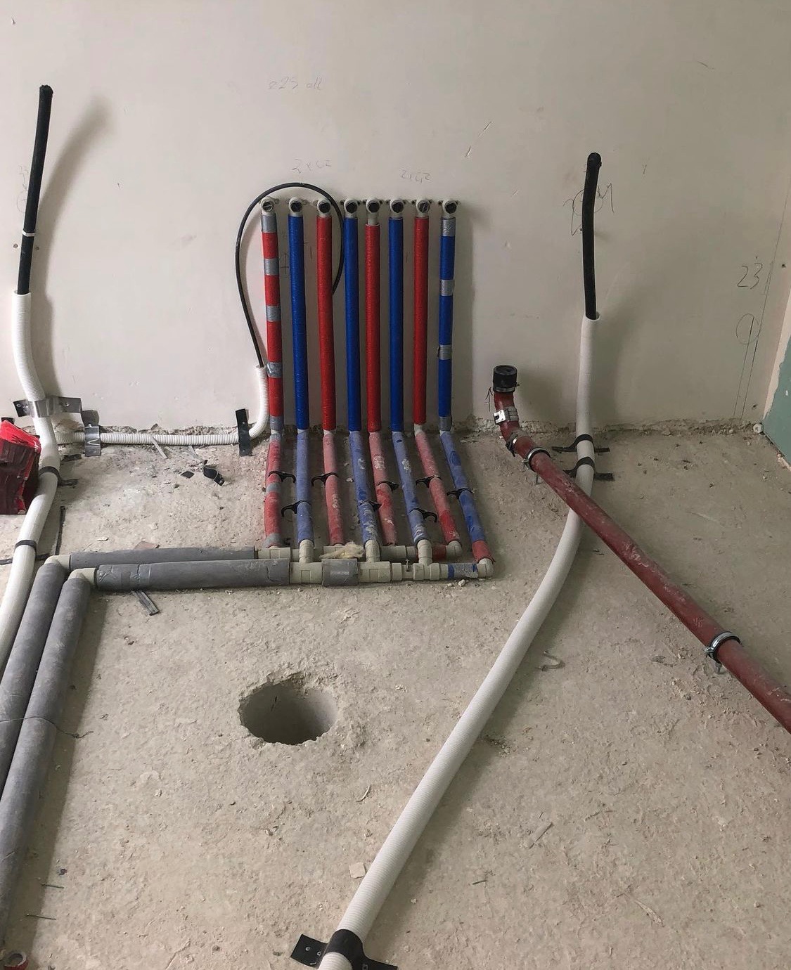 Wnętrze w trakcie montażu instalacji wodnej i grzewczej: widoczne pionowe rury w kolorach czerwonym i niebieskim przymocowane do ściany, poziome szare rury na podłodze oraz otwór w posadzce.