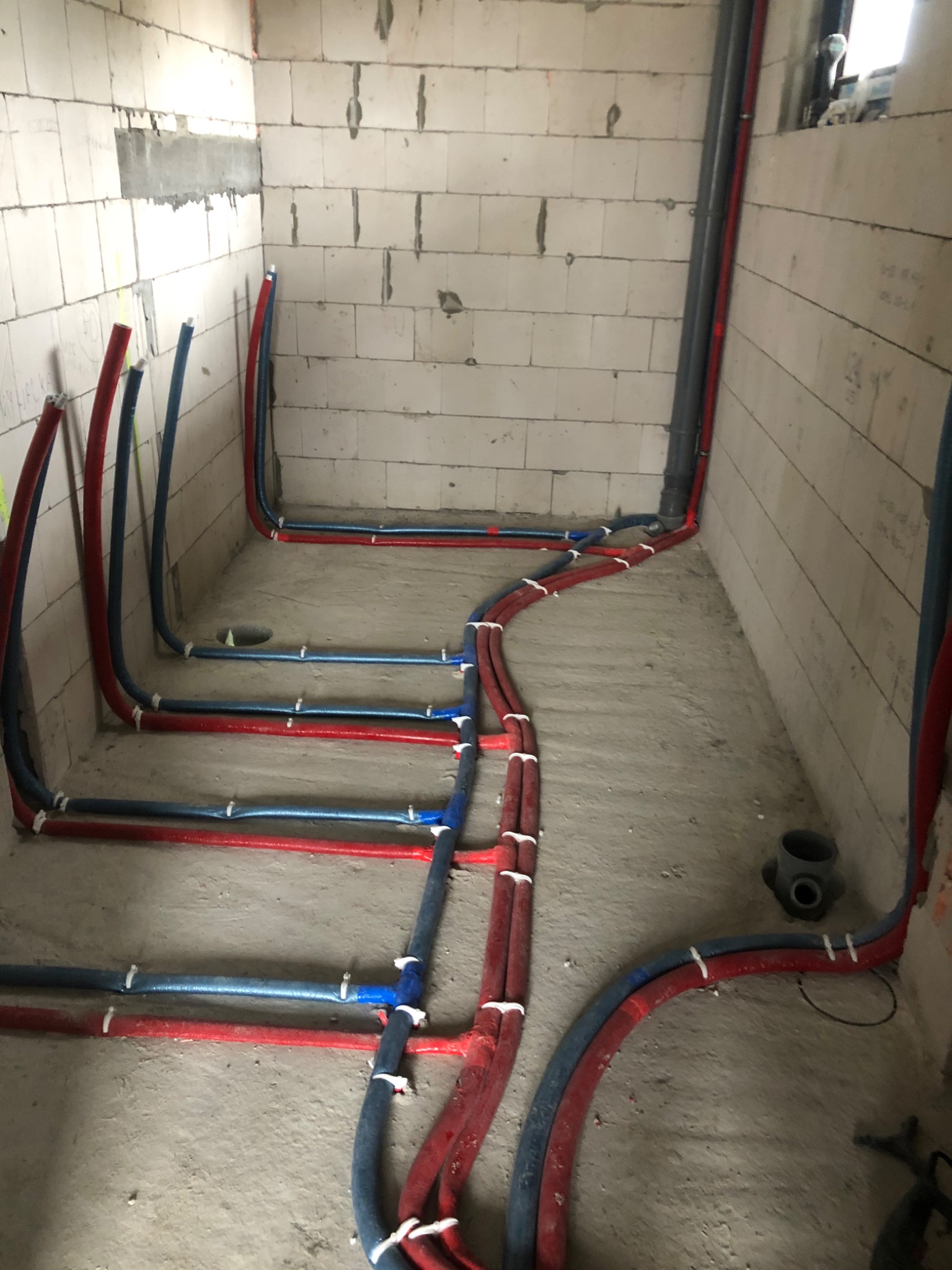 Wnętrze w trakcie instalacji hydraulicznej: czerwone i niebieskie rury prowadzone po podłodze i wzdłuż ścian z bloczków betonowych, mocowane białymi opaskami, widoczny trójnik kanalizacyjny.