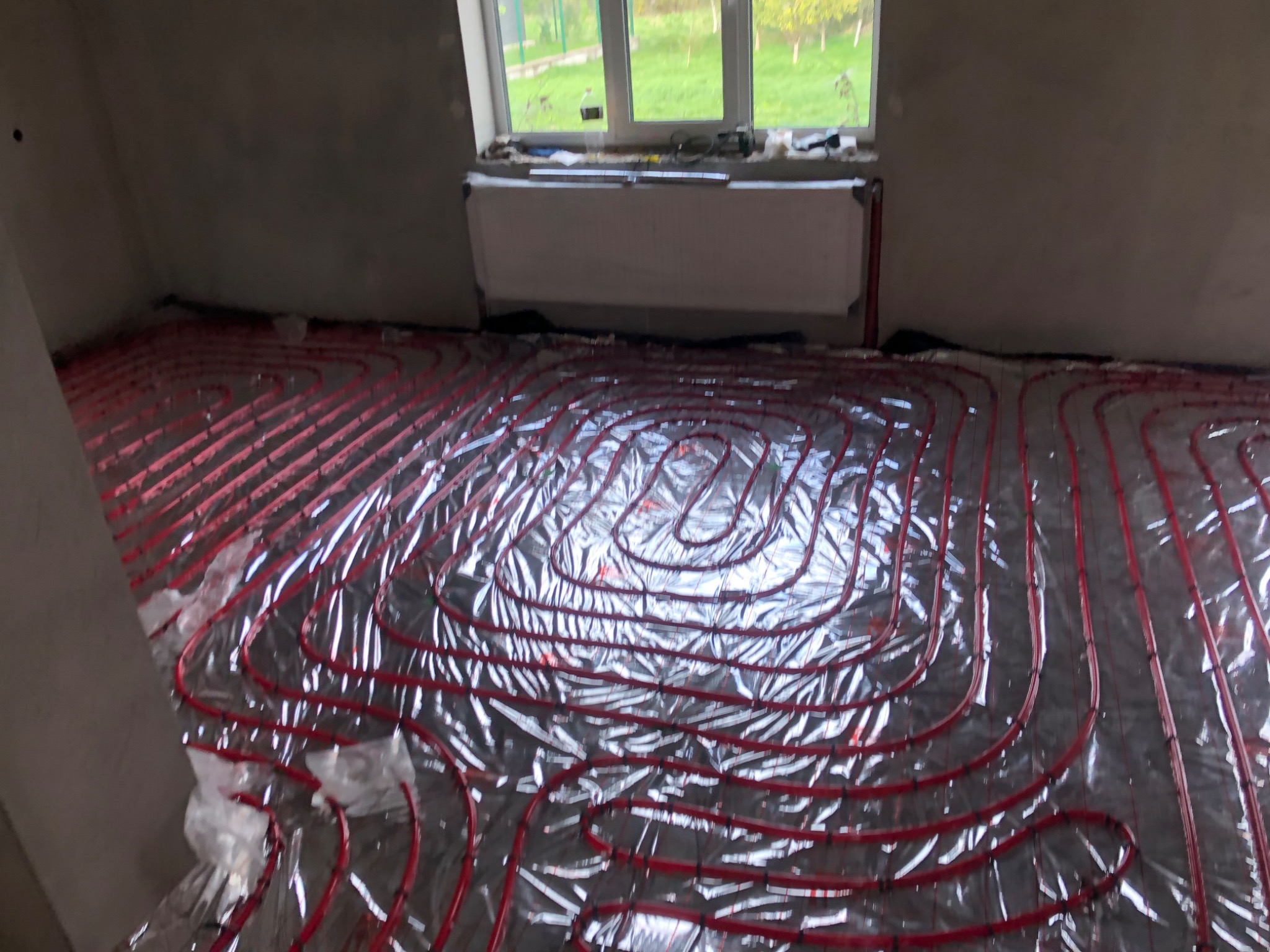 Czerwone rurki ogrzewania podłogowego ułożone na srebrnej folii izolacyjnej w surowym pomieszczeniu z grzejnikiem pod oknem.