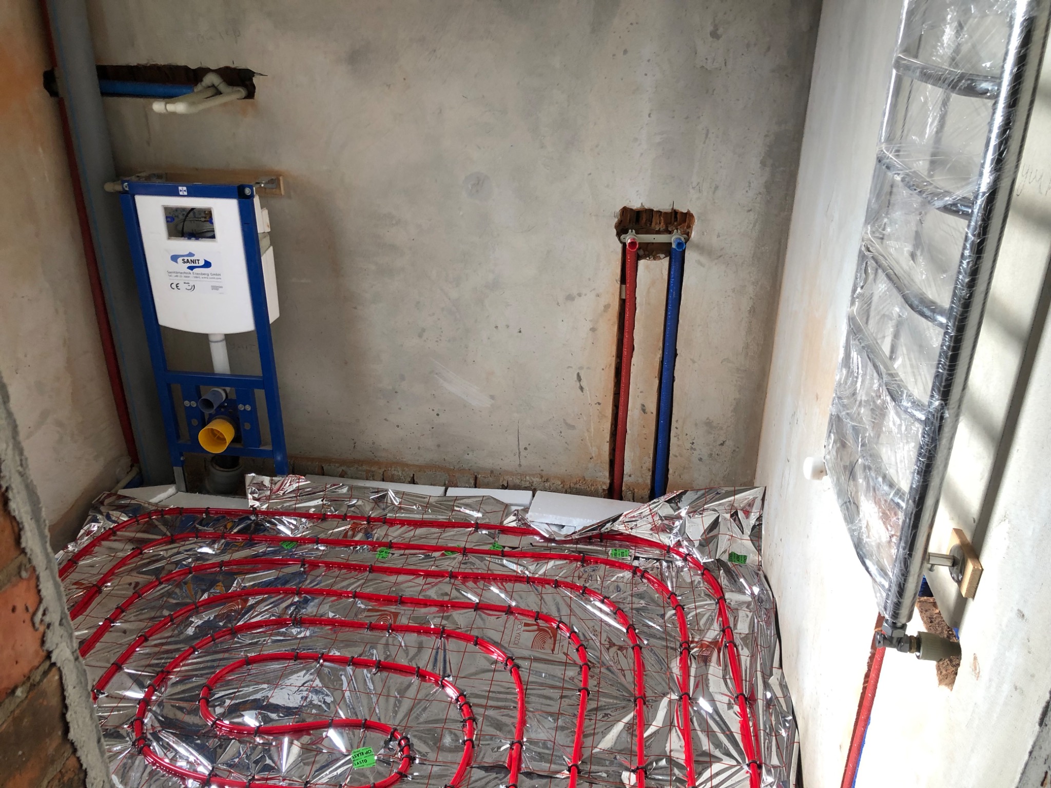 Wnętrze łazienki w trakcie remontu: widoczne czerwone rurki ogrzewania podłogowego ułożone na srebrnej folii, stelaż podtynkowy WC marki Sanit oraz grzejnik drabinkowy owinięty folią ochronną...
