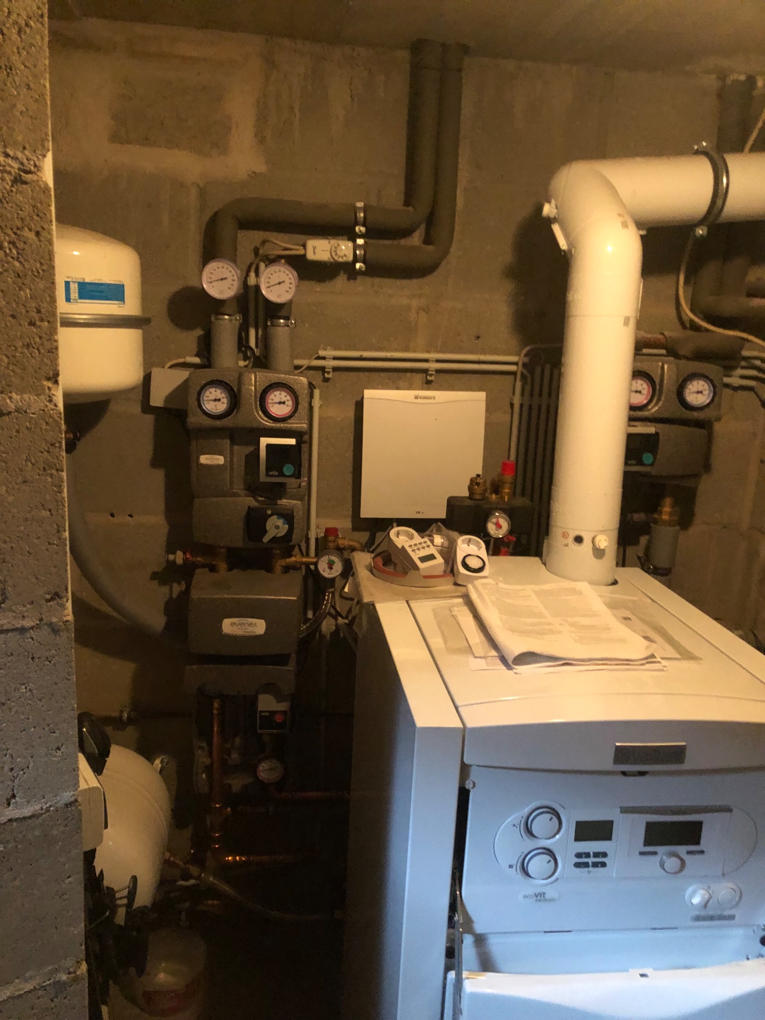 W kotłowni zainstalowany biały piec gazowy Vaillant ecoVIT, z widocznymi manometrami, rurami i elementami instalacji, na piecu leżą dokumenty, ściany z surowego betonu.