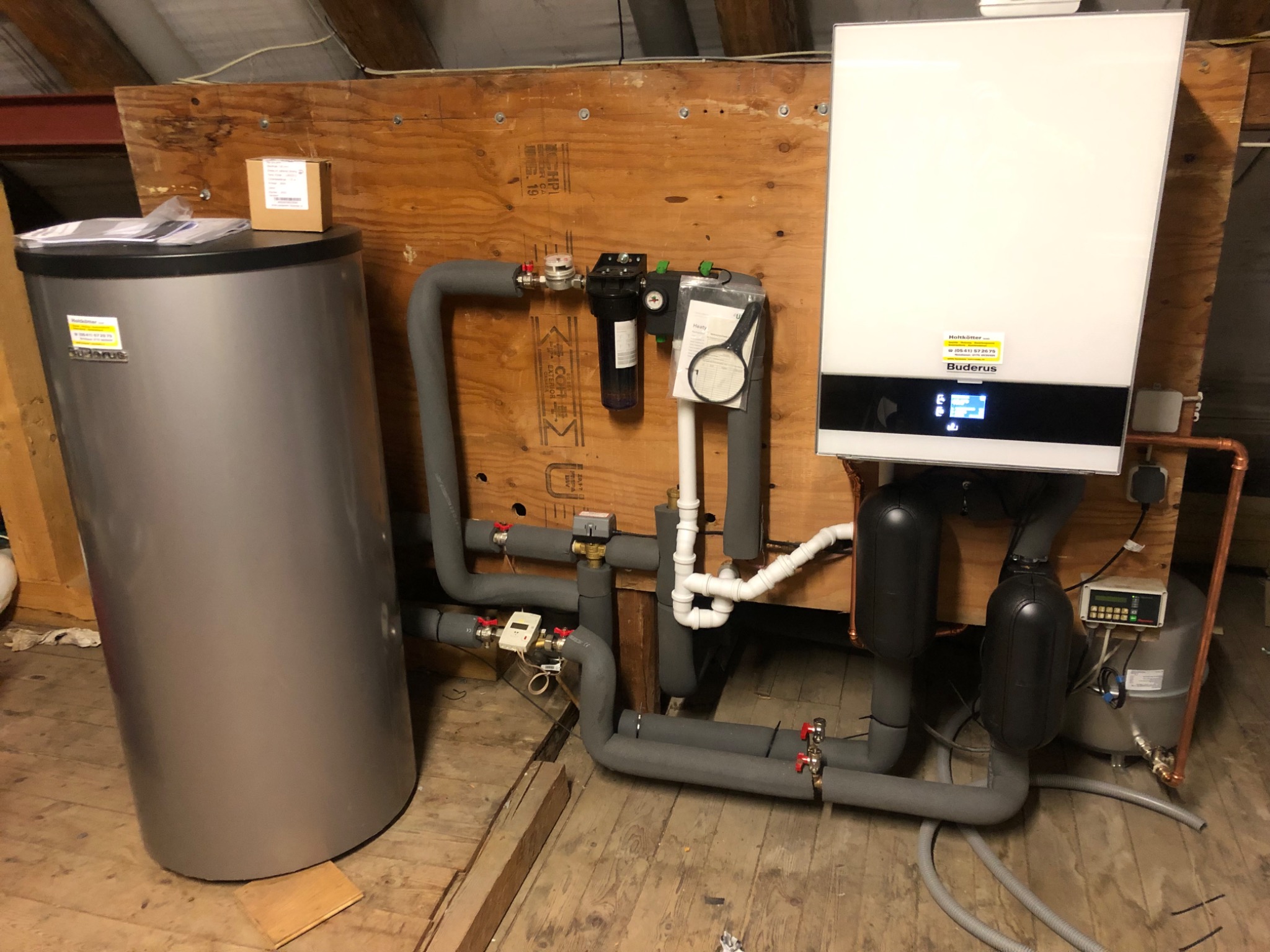 Nowoczesny, biały piec grzewczy Buderus z szarym zbiornikiem buforowym i rurami w izolacji, zamontowany na drewnianej ścianie w pomieszczeniu o drewnianej podłodze, widoczne liczniki i filtry...