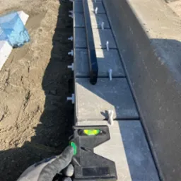 Układanie szarej kostki brukowej z użyciem poziomicy i plastikowych krzyżyków dystansowych, widoczny rów z ziemią i betonowy krawężnik.