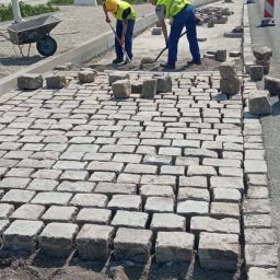ELIKA SPÓŁKA Z OGRANICZONĄ ODPOWIEDZIALNOŚCIĄ - Fragment układanej nawierzchni z kostki granitowej; widoczne narzędzia i pracownicy w odblaskowych kamizelkach; w tle droga i chodnik z obrzeżami.