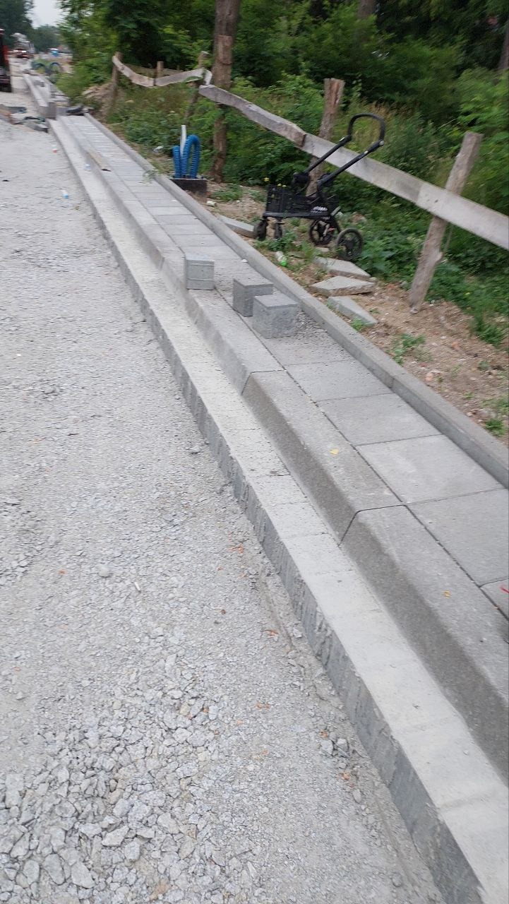 Montaż obrzeży chodnika z kostki betonowej, widoczne niedokończone fragmenty i przygotowane kostki, w tle fragment wózka transportowego.