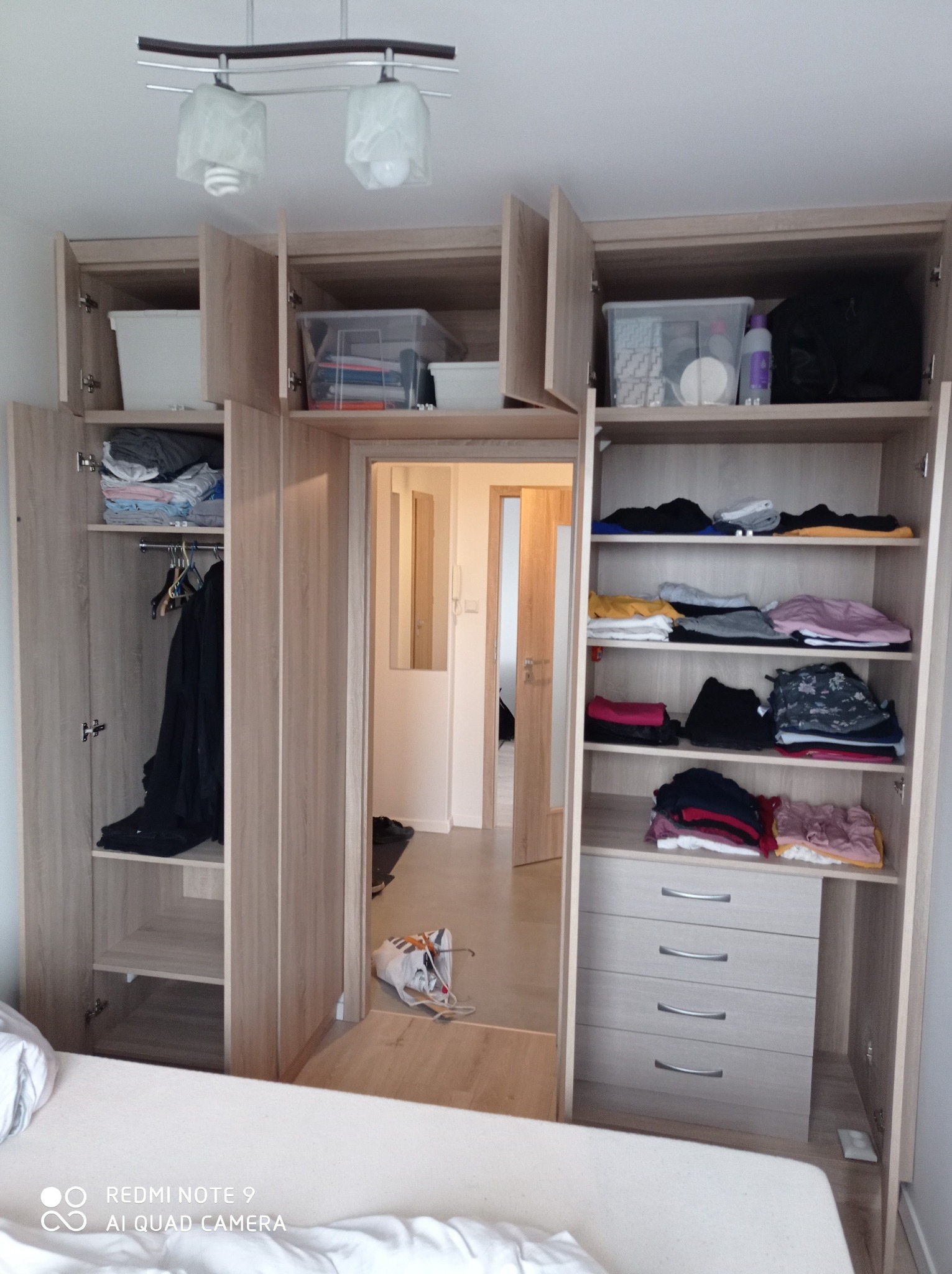 Jasna, drewnopodobna szafa wnękowa z otwartymi drzwiami, ukazująca półki z ubraniami, szuflady i przejście do kolejnego pomieszczenia.