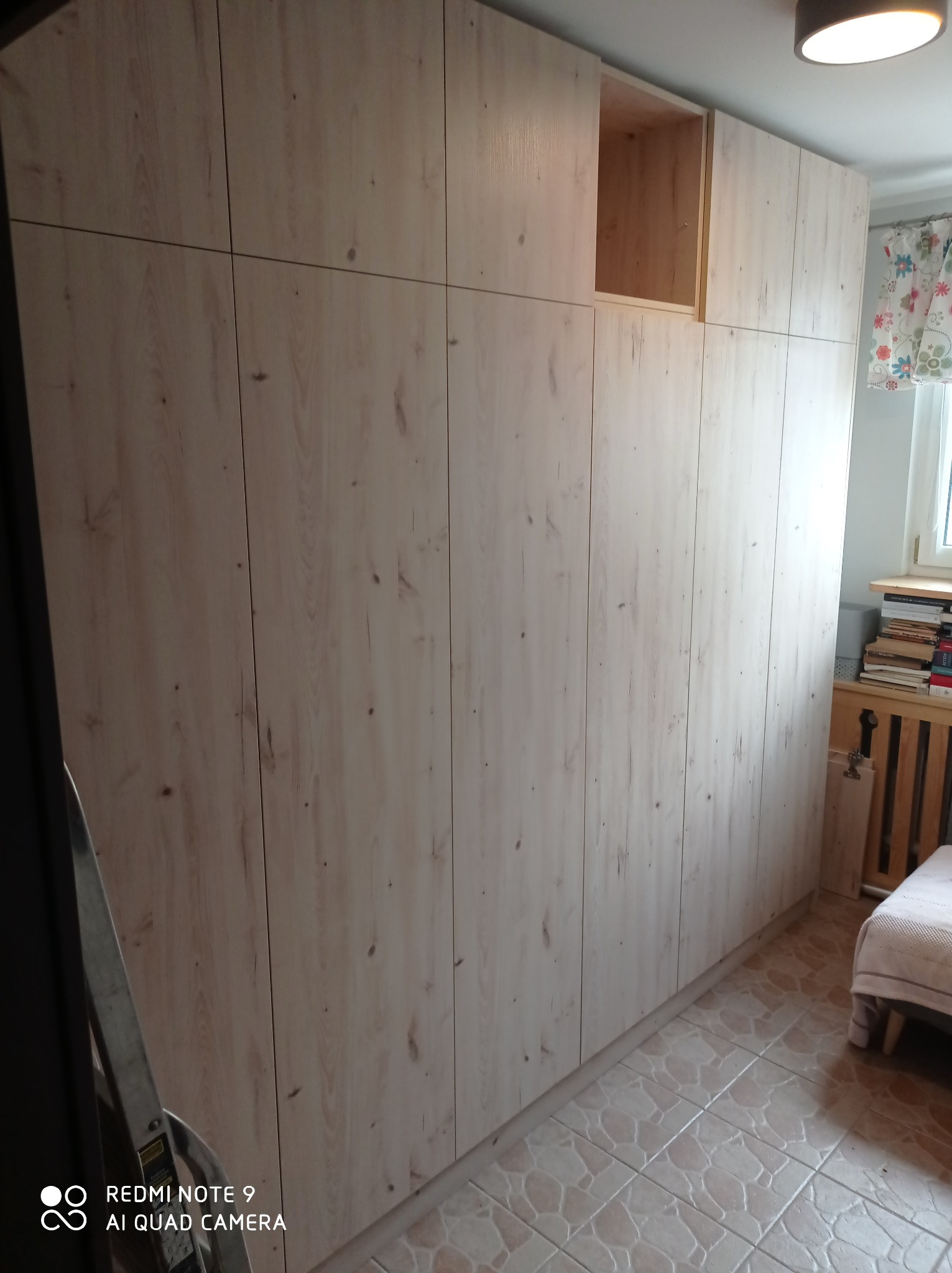 Wysoka zabudowa meblowa z jasnego drewna z widoczną strukturą słojów, obejmująca szafki górne i dolne, z otwartą przestrzenią w górnej sekcji, na tle pokoju z oknem i grzejnikiem.