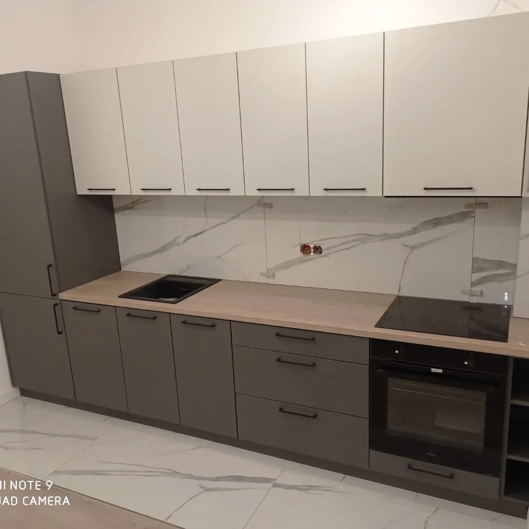 Nowoczesna zabudowa kuchenna z szarymi dolnymi szafkami i białymi górnymi, czarnymi uchwytami i blatem imitującym drewno, z marmurowym panelem nad blatem.