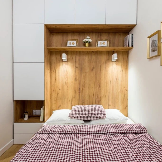 Nowoczesna sypialnia z zabudową meblową: białe szafki, drewniana wnęka z półkami oświetlonymi kinkietami, łóżko z bordowo-białą pościelą w pepitkę.