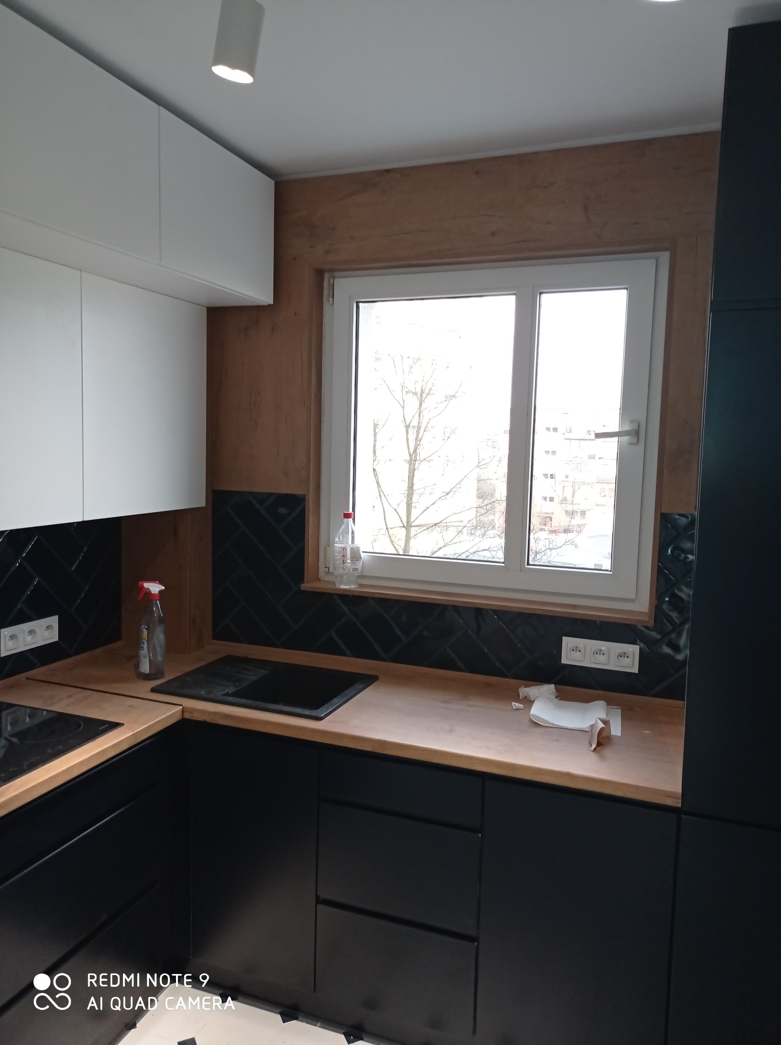 Nowoczesna kuchnia w Bydgoszczy z czarnymi frontami szafek, drewnianym blatem i czarnymi płytkami ułożonymi w jodełkę nad blatem. Widoczne białe szafki wiszące i okno z widokiem na zabudowę miejską.