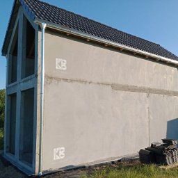 Prefa Invest Sp. z o.o. - Surowy stan dwukondygnacyjnego domu murowanego z widoczną konstrukcją i zadaszeniem, przygotowanego do dalszych prac wykończeniowych, z logo firmy na elewacji i stosem dachówek obok.