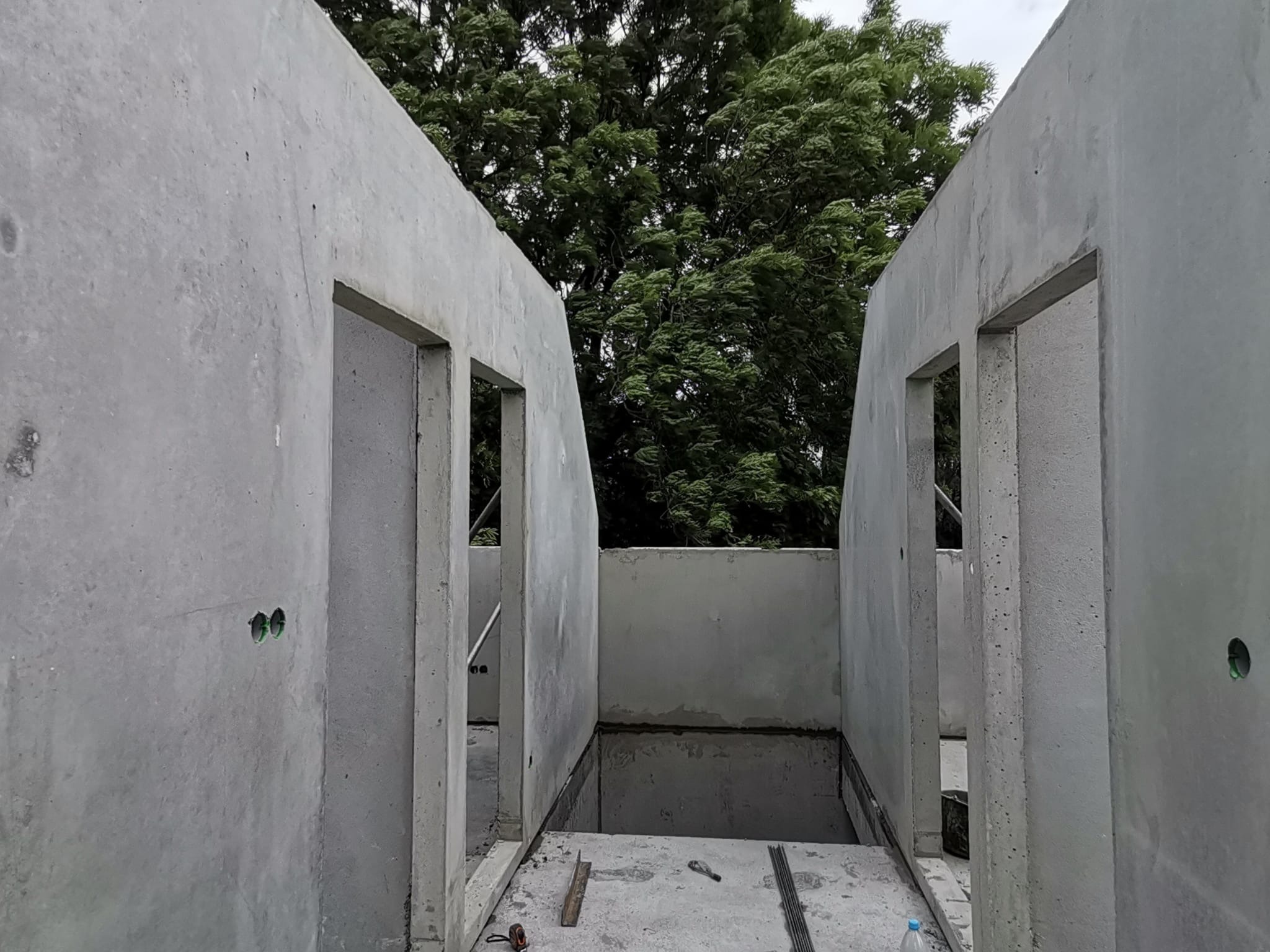 Surowe, betonowe ściany z otworami okiennymi i drzwiowymi w trakcie montażu, widoczne elementy zbrojenia i narzędzia na posadzce, drzewa w tle.