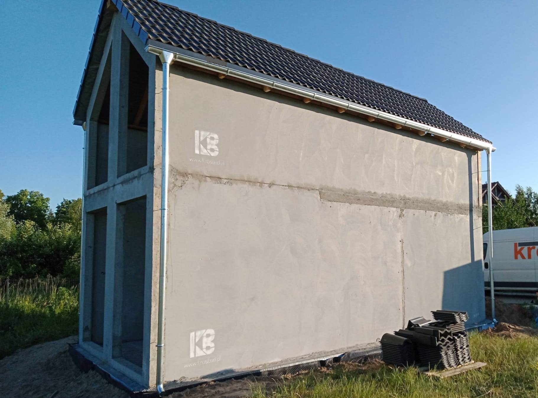 Surowy stan dwukondygnacyjnego domu murowanego z widoczną konstrukcją i zadaszeniem, przygotowanego do dalszych prac wykończeniowych, z logo firmy na elewacji i stosem dachówek obok.