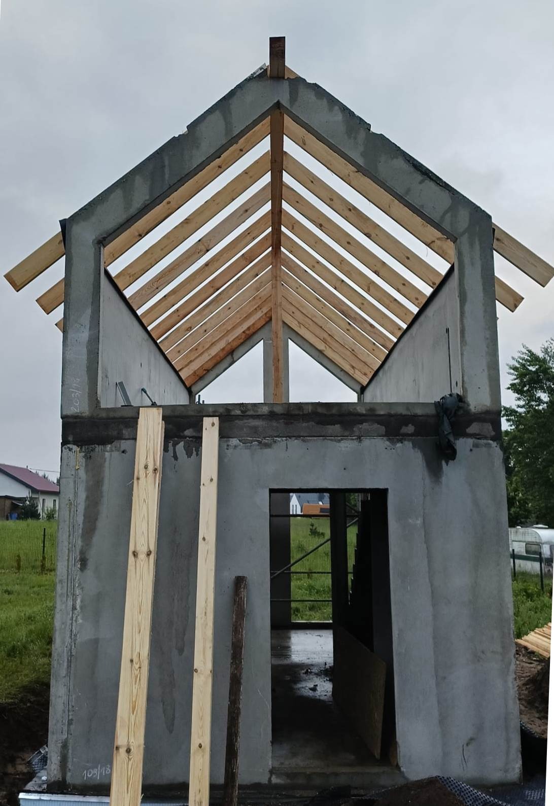 Surowy stan domu w trakcie budowy: betonowe ściany, drewniane elementy konstrukcji dachu, widoczne deskowanie i zarys otworu drzwiowego.