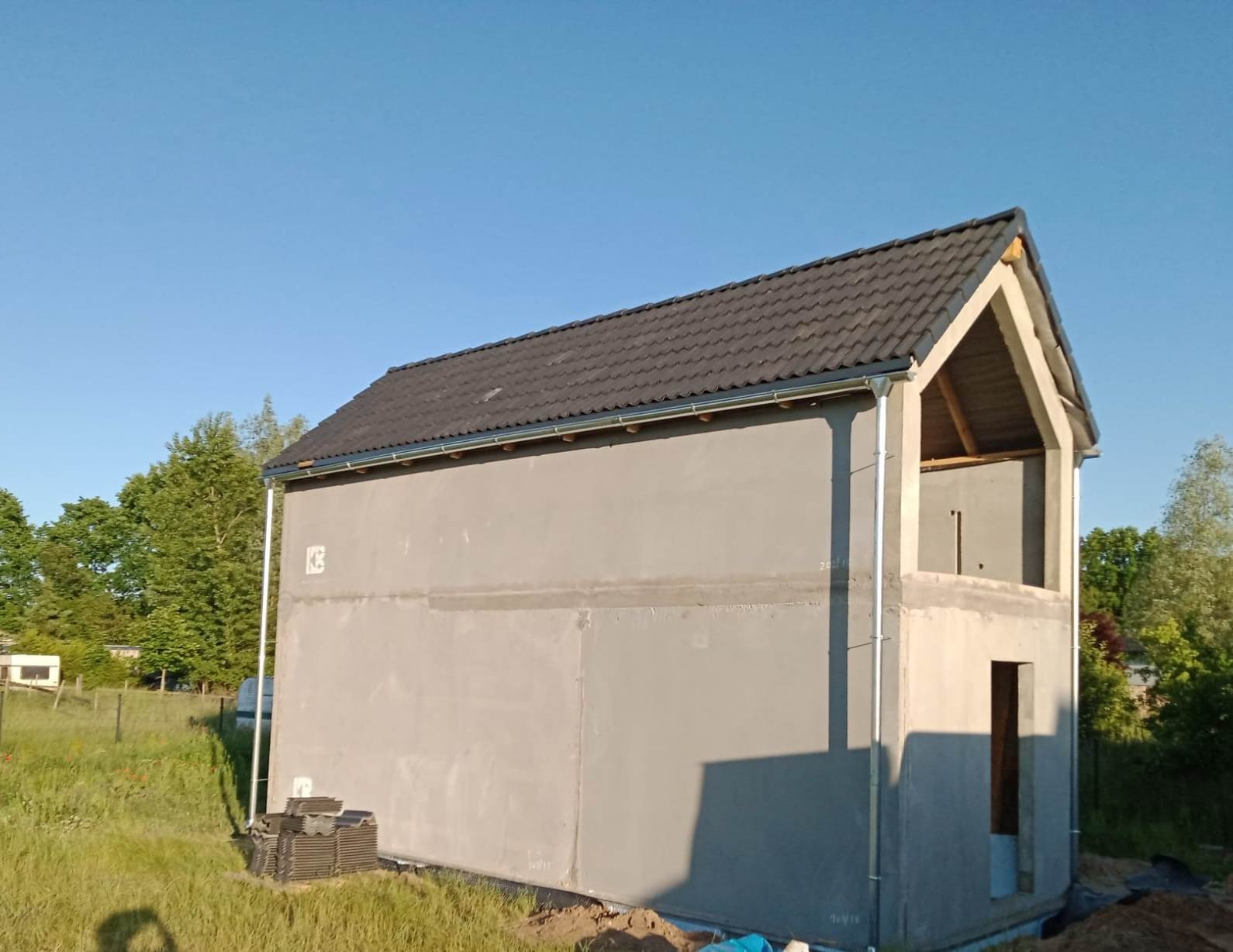 Surowy, dwukondygnacyjny budynek w trakcie budowy z szarą elewacją, czarnym dachem i widoczną drewnianą konstrukcją poddasza, otoczony zielenią i błękitnym niebem.