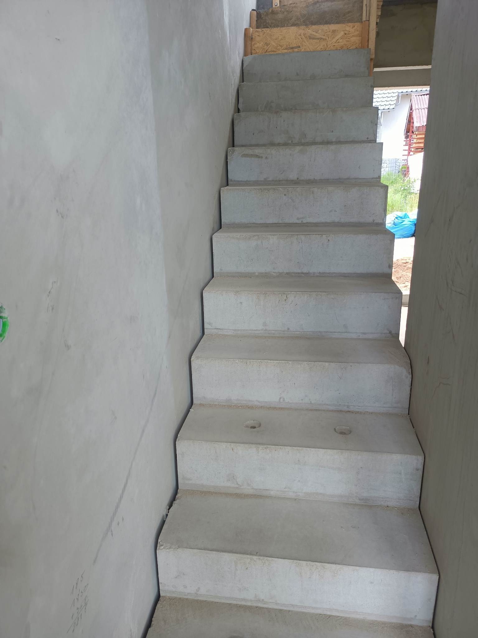 Surowe betonowe schody wewnętrzne w trakcie budowy domu, widoczne ślady po deskowaniu i otwory technologiczne w stopniach, ściany w stanie surowym.