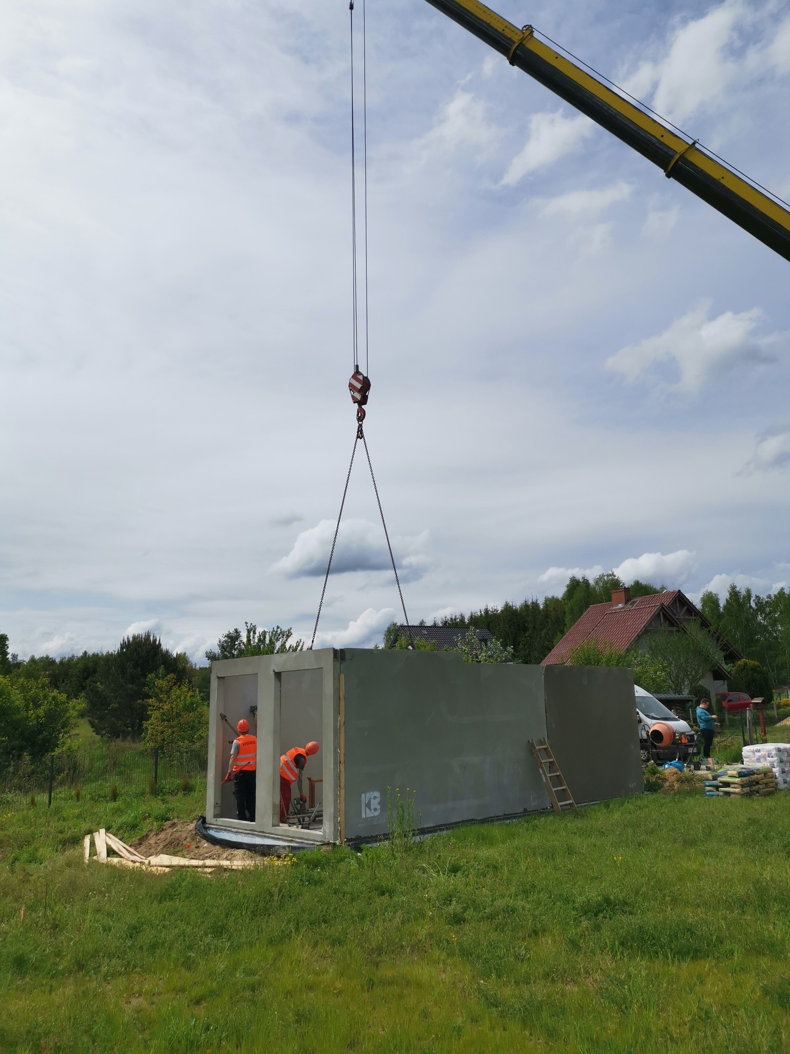 Montaż modułu betonowego domu przy użyciu dźwigu, dwóch pracowników w pomarańczowych kamizelkach wewnątrz, widoczne worki z zaprawą i betoniarka w tle.