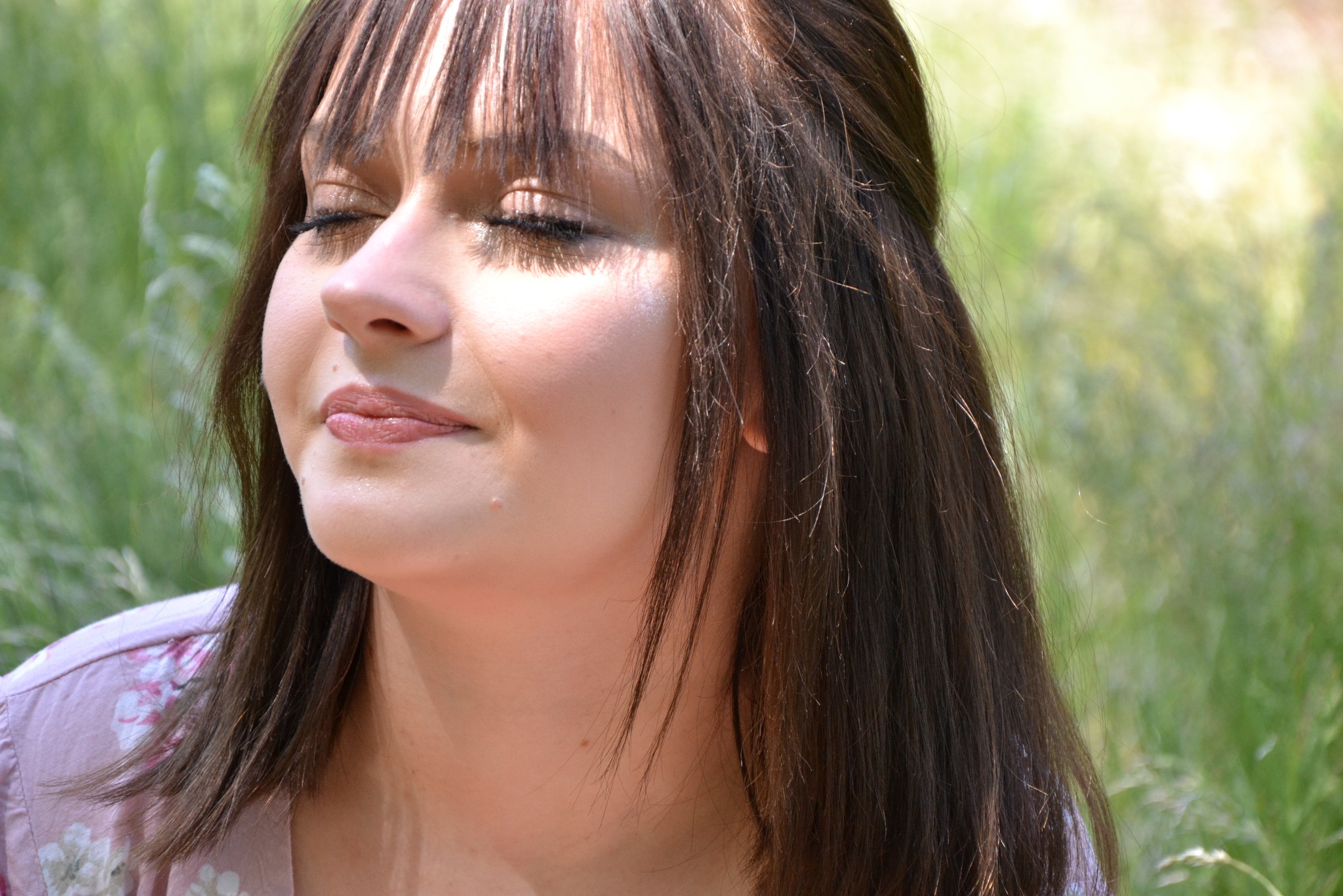 Portret kobiety z zamkniętymi oczami, delikatny makijaż, brązowe włosy z grzywką, rozproszone światło, tło z zielenią.