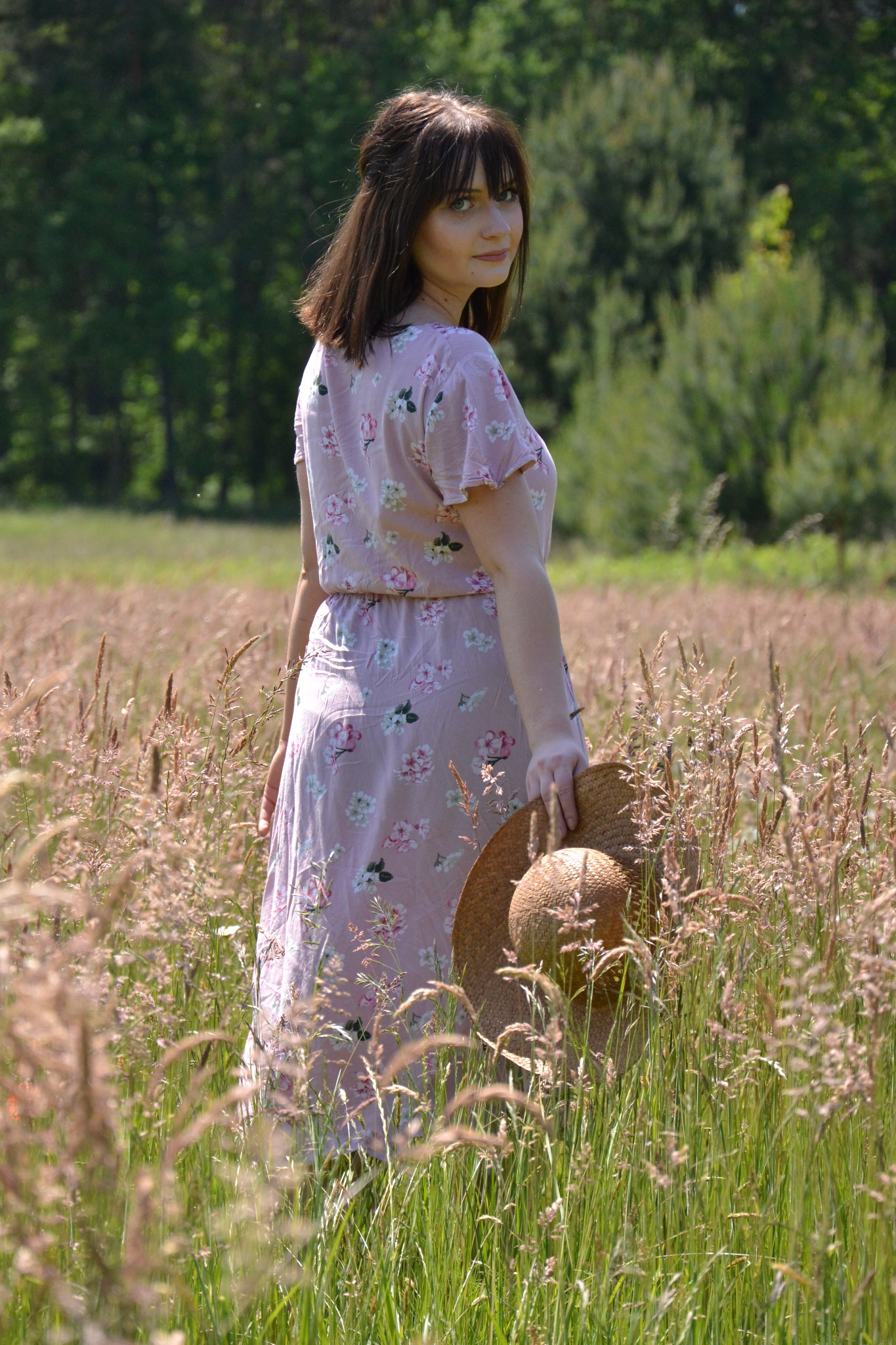 Kobieta w kwiecistej sukience i kapeluszem na łące w Szczedrzyku, odwrócona przez ramię z delikatnym uśmiechem.