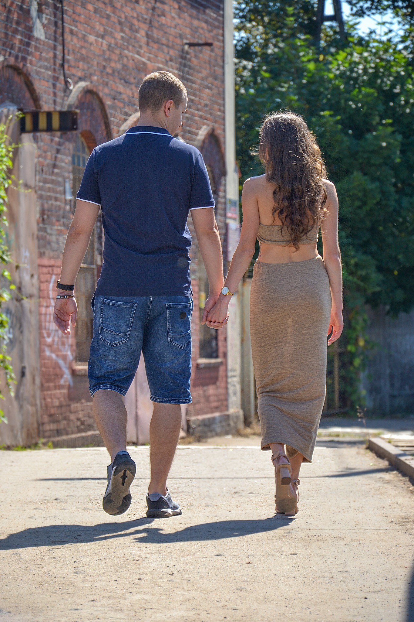 Para idzie ulicą trzymając się za ręce, widok z tyłu, kobieta w dopasowanym stroju i mężczyzna w krótkich spodenkach, ceglany budynek w tle.