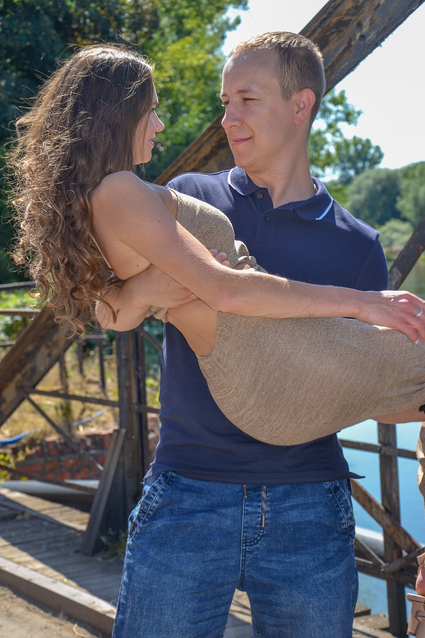 Mężczyzna w granatowej koszulce polo i jeansach trzyma na rękach kobietę w beżowej sukience na tle zieleni i konstrukcji mostu.