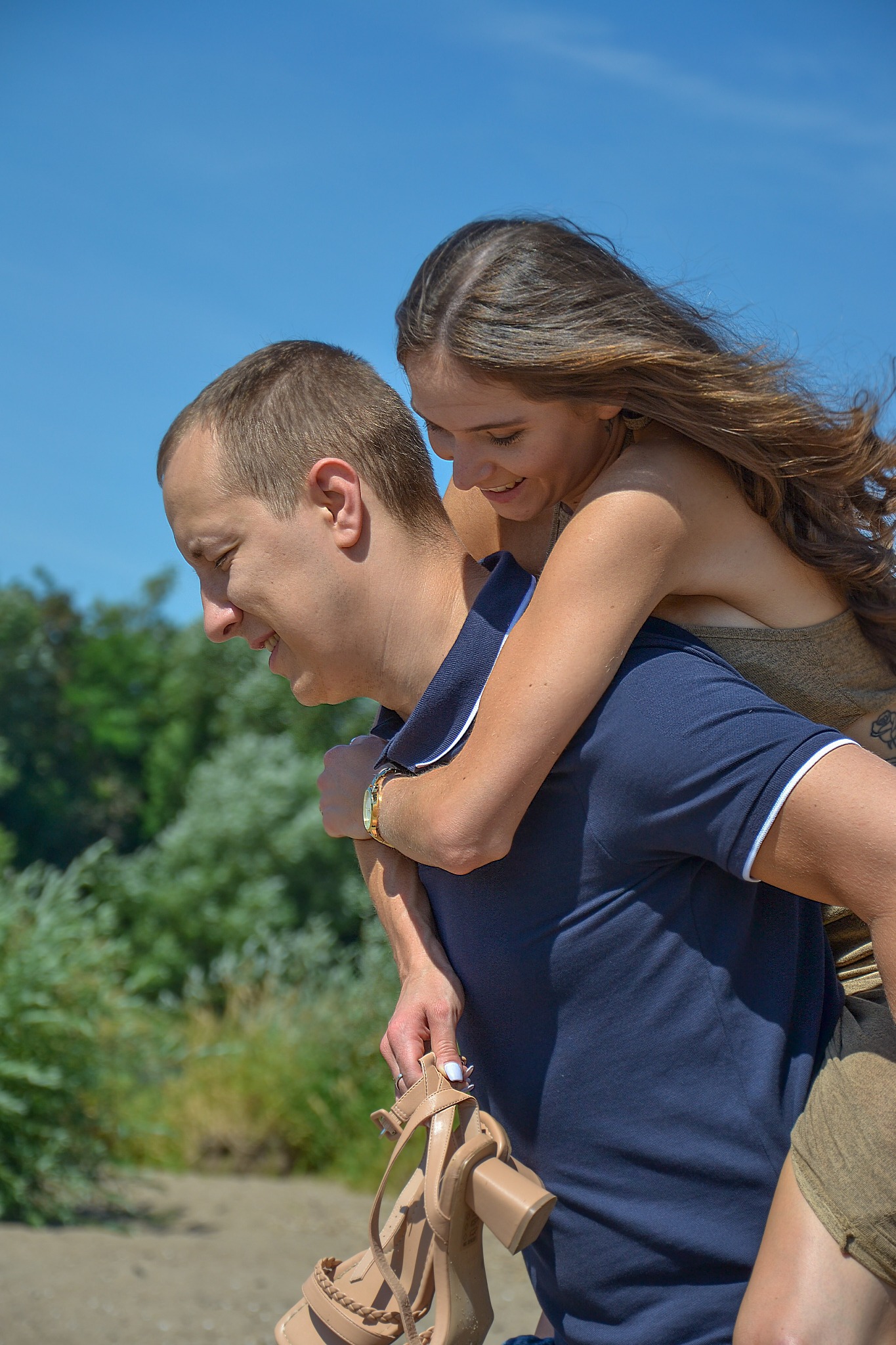 Uśmiechnięta kobieta na barana u mężczyzny, trzymająca beżowe sandały na tle zieleni i błękitnego nieba.