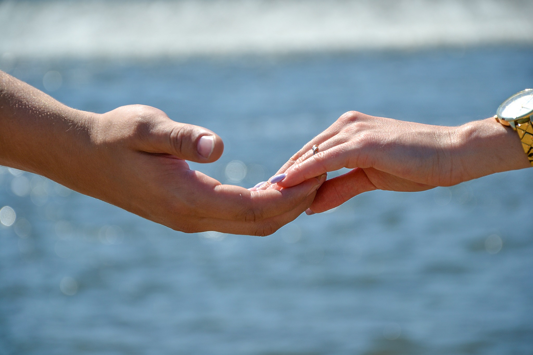 Zbliżenie dłoni kobiety z pierścionkiem zaręczynowym, dotykającej dłoni mężczyzny na tle rozmytej wody w Szczedrzyku, złoty zegarek na damskim nadgarstku.
