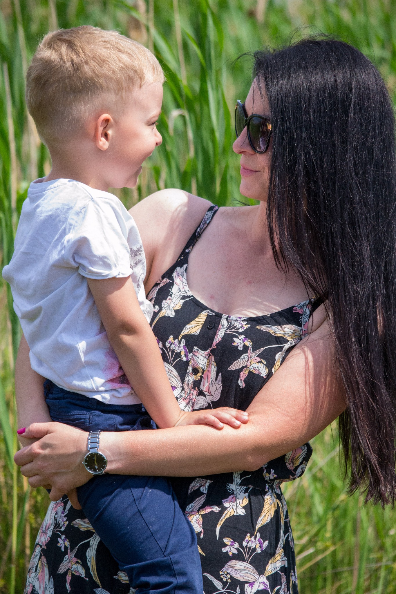 Kobieta w kwiecistej sukience i okularach przeciwsłonecznych trzyma na rękach jasnowłosego chłopca na tle zielonych traw.