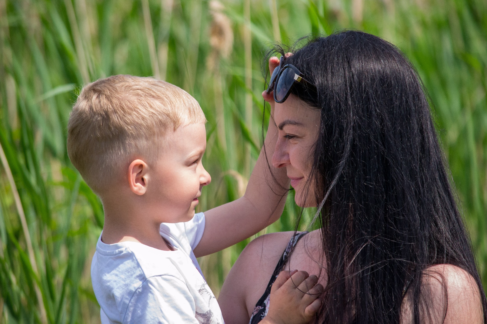 Profil matki z czarnymi włosami i okularami na głowie, obejmującej blond synka na tle zielonych traw.