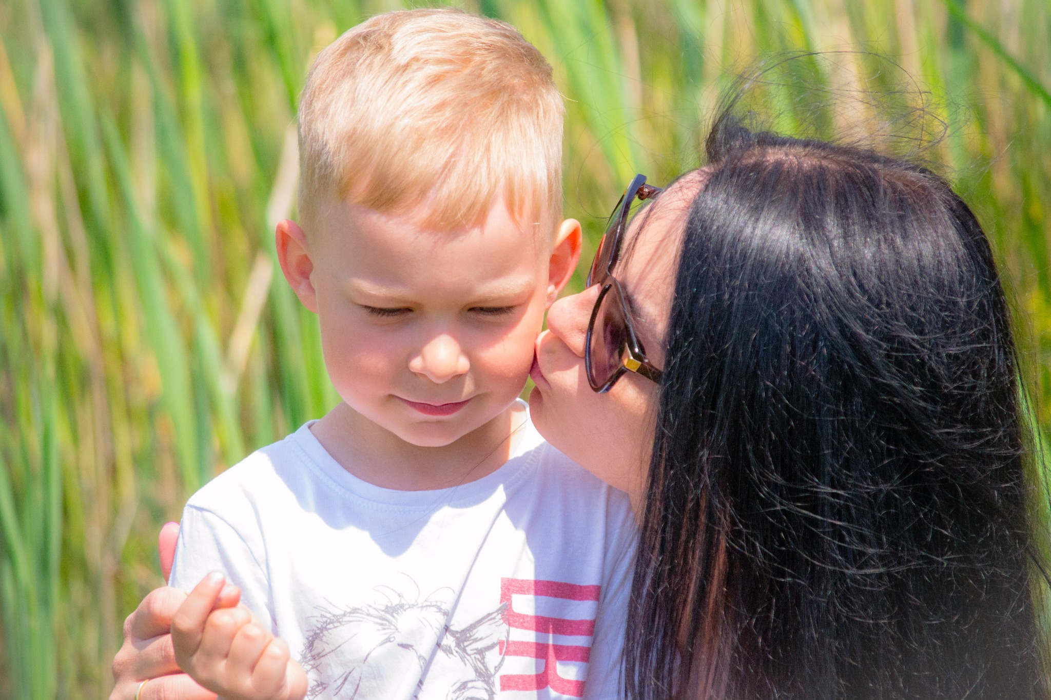 Matka w ciemnych okularach całuje w policzek blond chłopca na tle zielonych traw.