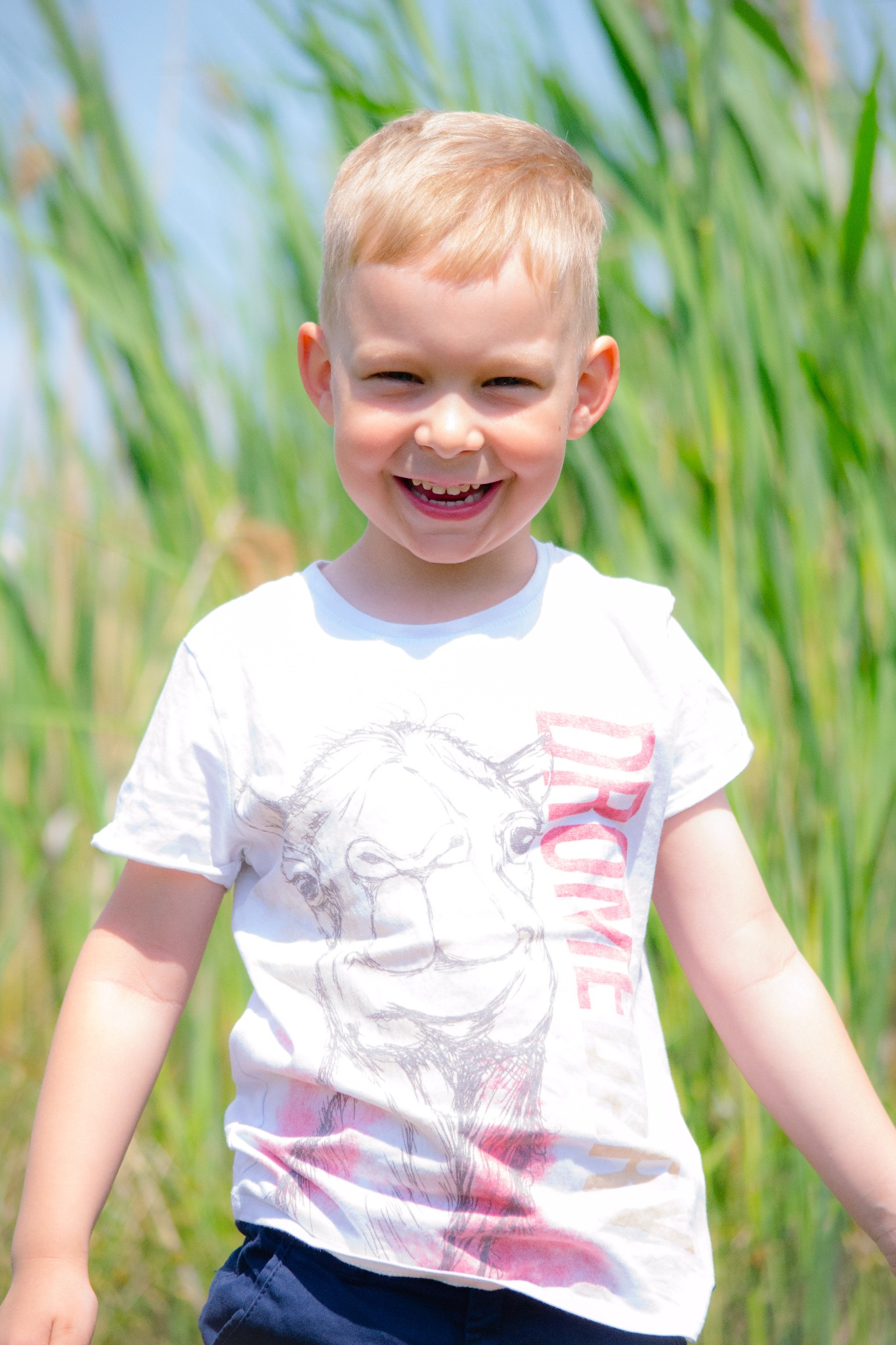 Uśmiechnięty blond chłopiec w białej koszulce z nadrukiem, na tle zielonych traw i błękitnego nieba.