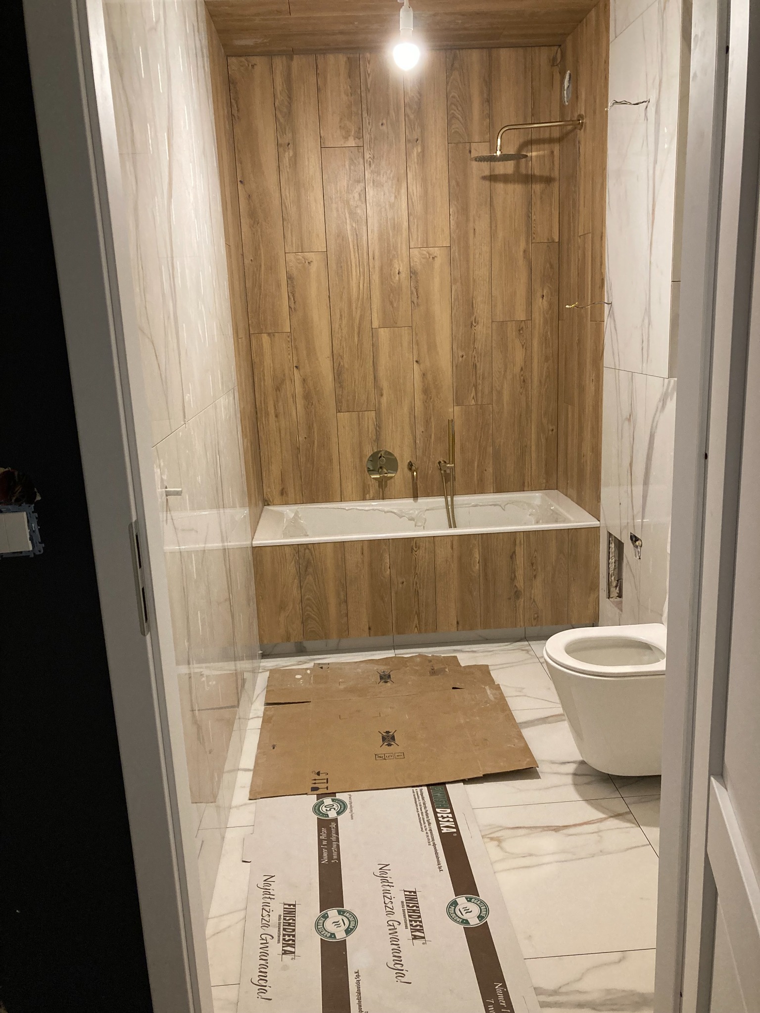 Łazienka w trakcie wykańczania, z wanną obudowaną drewnopodobnymi panelami, złotą armaturą i częściowo ułożonymi marmurowymi płytkami, zabezpieczonymi kartonem i folią ochronną.