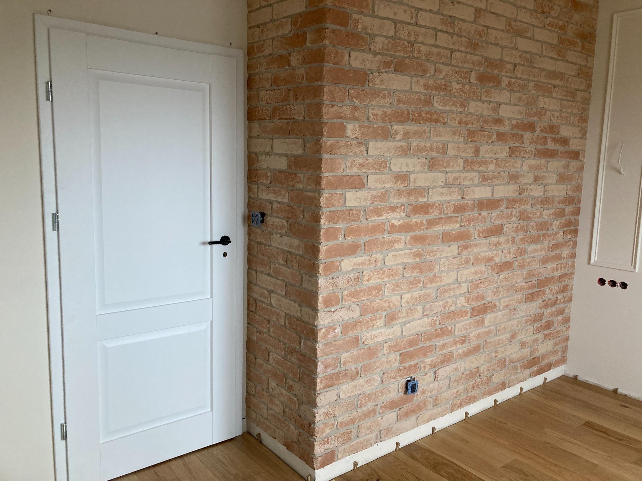 Ściana wykończona cegłą dekoracyjną w odcieniach czerwieni i beżu, z widoczną białą stolarką drzwiową i fragmentem drewnianej podłogi, gniazdka elektryczne niezamaskowane.